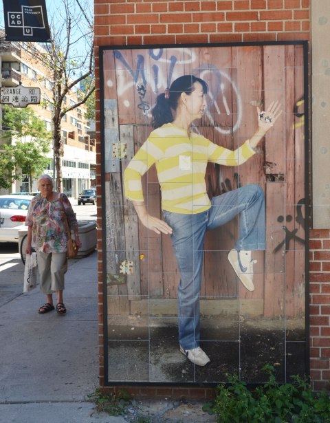 A picture of a woman in profile, with one knee raised, wearing a yellow and white striped top, picture on a wall. An older woman is walking on the sidewalk, approaching the camera. 