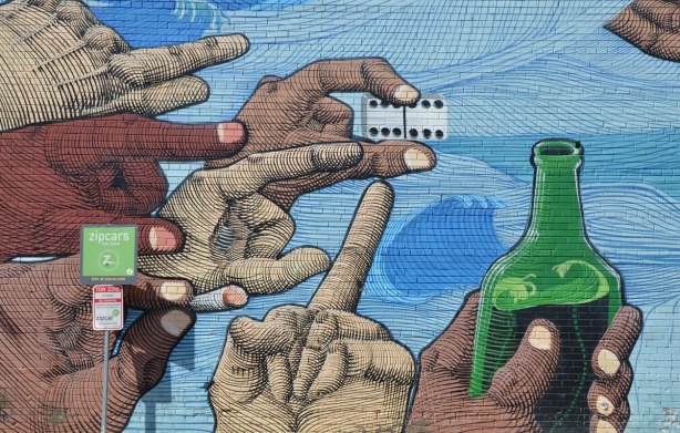 close up of bottom left of a mural showing 7 hands of different shades of brown and beige. One is holding a pair of dice (two sixes), one is holding a green bottle, presumably with alcohol in it, the other fingers are pointing to the right, towards the main part of the mural. 