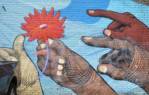 part of a mural by Nunca, four hands of differing shades of brown and beige are pointing or holding a large orange flower