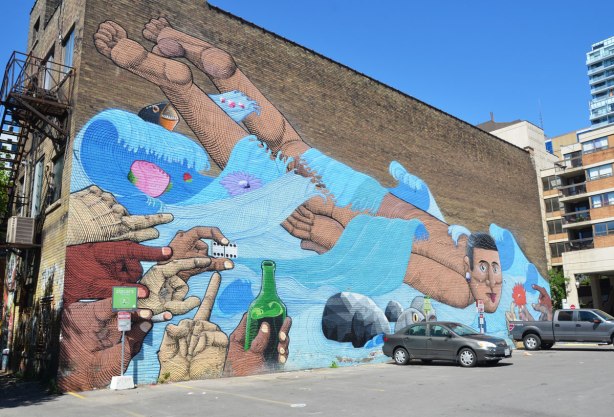 large mural on an exterior wall beside a parking lot. A man is swimming away from hands holding booze and dice and towards hands holding flowers. There are a few fish in the water with him. Painted by street artist Nunca on a bulding on McCaul Street in Toronto 