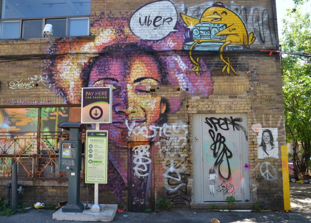 street art piece of a woman's face with eyes closed and a vague shape of a heart behind her, done in purples and yellows, by Dasic Fernandez, on the side of brick building, with a pay machine for a parking lot in front of it. Just above her is an UBER5000 birdie with a ghetto blaster