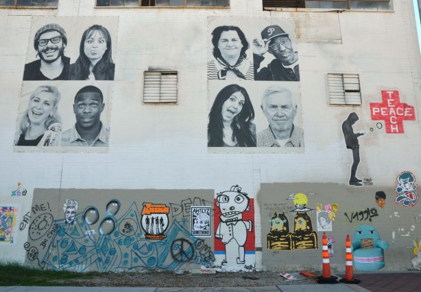 eight large black and white photos of people from the shoulders up on the upper level of a building wall. On the lower level is some graffiti and street art. 