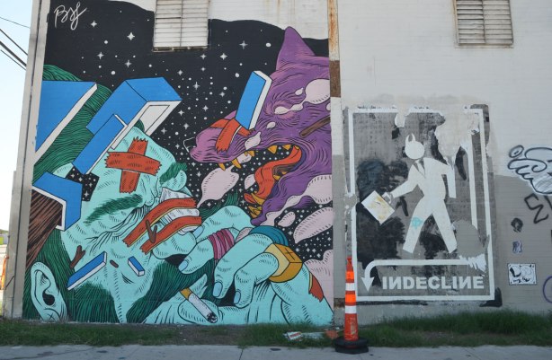 a mural and a wheatpaste. The mural is a green and light blue man's head, fingers by his face, cigarette in his mouth. A purple dog is lunging at him with its mouth open. The wheatpaste is a silhouette man carrying a briefcase with the word indecline written below him. 