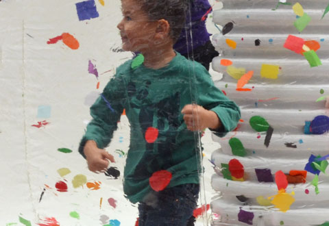 A boy stands inside a large clear plastic bubble with many bits of coloured paper. He is batting at the papers and smiling. 