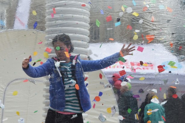 A woman stands inside a large clear plastic bubble with many bits of coloured paper. She is posing for a photograph. 