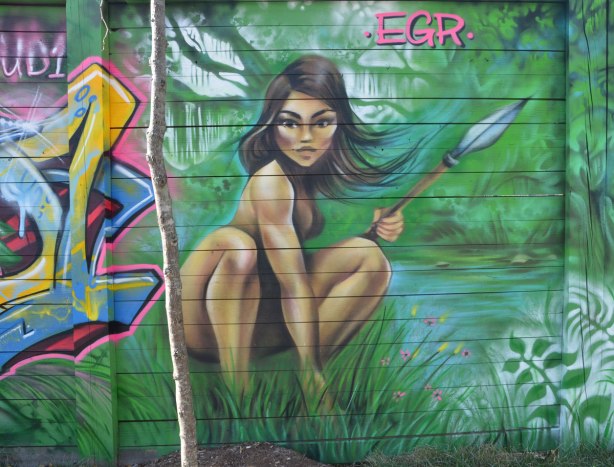 street art mural painting by EGR of a probably naked woman crouching in the long grass with a spear in her hand. 