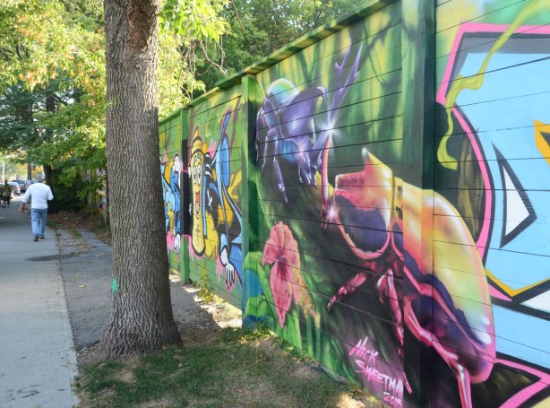 side view of a street art mural on a wall beside a sidewalk, the closest part of the mural is a picture of two giant beetles by Nick Sweetman with a painting of a spray paint can farther down the fence. 