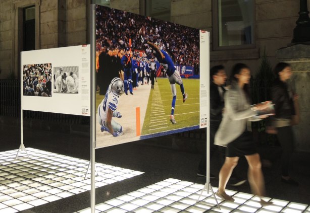 Picture taken at night. The light source is from lights in the floor. Three photographs are on display, part of a larger exhibit of winning photography from around the world. The three shown here are sports photos. The main one being a football player catching a pass. 