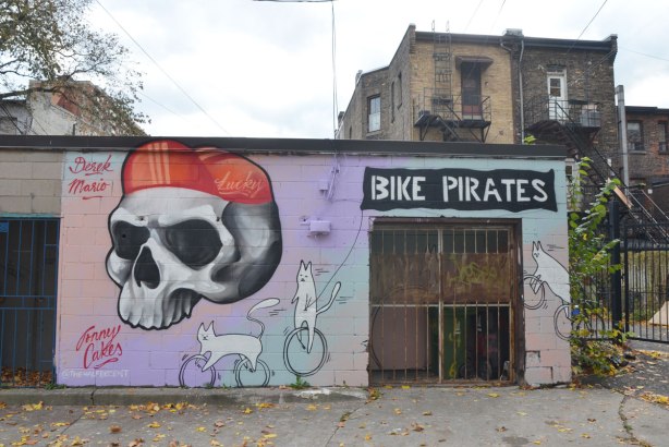 In a laneway, painted by Jonny Cakes @thehalfdecent, the wall of a workshop, shed or garage, is painted with a big skull wearing a red bike hat. Two white cats, one on a unicycle and one on a bicycle, and the words BIKE PIRATE written over the door. 