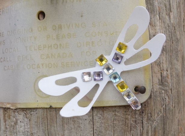 Small graffiti piece of a paper cut out dragonfly with sequins glued onto it's body and a small part of the wing. 