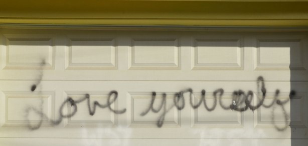 scrawled in cursive writing with black spray paint on a white garage door are the words love yourself