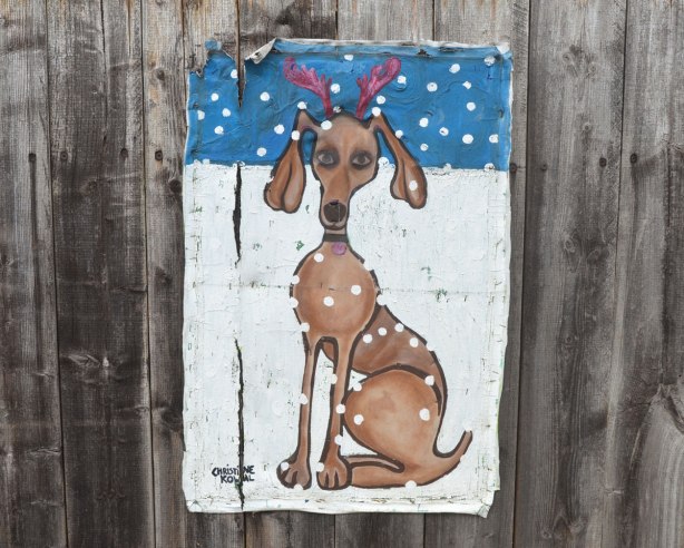 Painting on canvas stapled to a wood fence. A large dog is sitting in the snow. Red fake reindeer antlers on its head. More snow is falling from the sky.