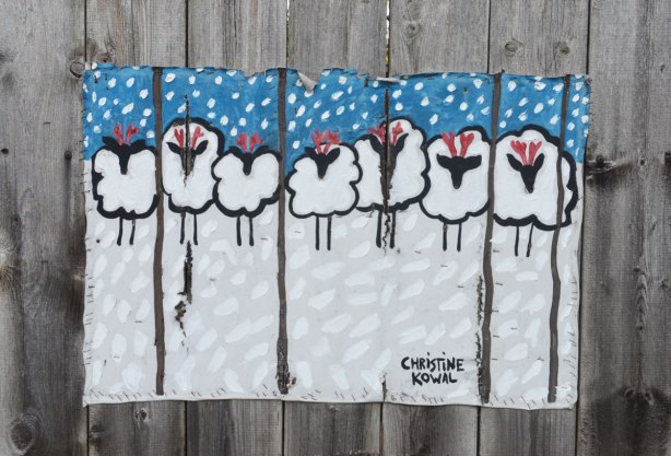 A painting of seven sheep in a line in the snow with more snow falling from the sky. The painting is on canvas and it is stapled to the fence.