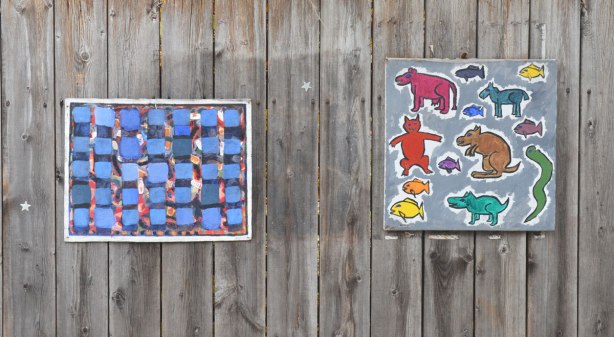 Two paintings on a wood fence. One is a collection of animals in bright colours and the other is an abstract with a large amount of blue in it.