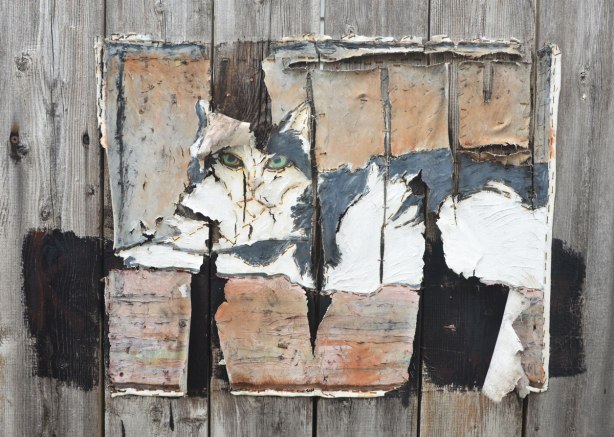 A very weathered painting of a cat on canvas and stapled to a wood fence. The canvas has started to rip vertically in places. The pattern of the rips sort of matched the pattern of the boards in the fence.