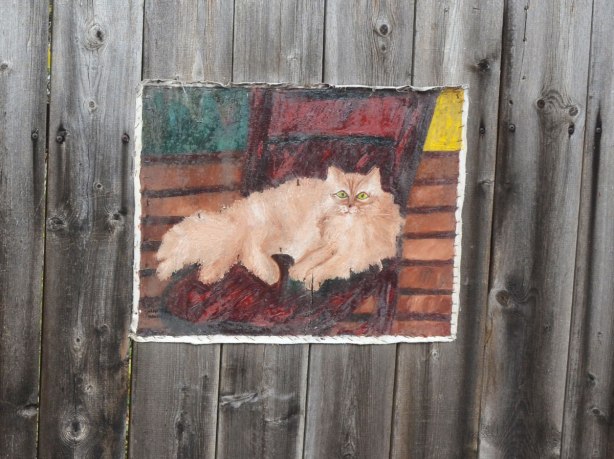 Painting on canvas stapled to a wood fence. A long haired light brown cat resting on a striped carpet or blanket