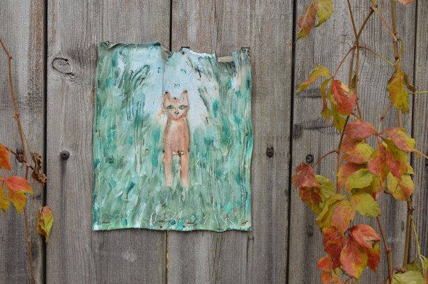 Painting on canvas stapled to a wood fence. A small brown cat in amongst tall grass. A vine is growing on the fence beside the picture.