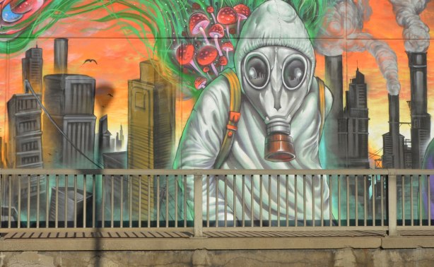 part of a mural by Essencia Art Collective on Lawrence West in Toronto showing the drastic possible environmental effects of not looking after the planet - a person covered in a grey suit and wearing a gas mask in front of a decaying and falling down city