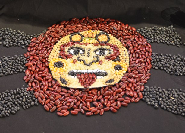 a face shape made of dried beans and corn. red beans make a circle around the face, black beans make 6 rays coming out from the circle. The face is corn with bean features. 