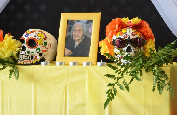 an ofrenda with a picture of a woman in a frame sitting on a table. One each side of her is an elaborately decorated skull. One of the skulls is wearing sunglasses and a wreath of yellow and orange flowers around the top of its head. 