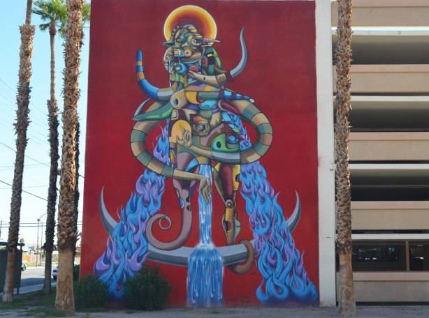 A large creature mural, yellow and orange halo around its head, blue furry legs, 5 storeys high, red background. 