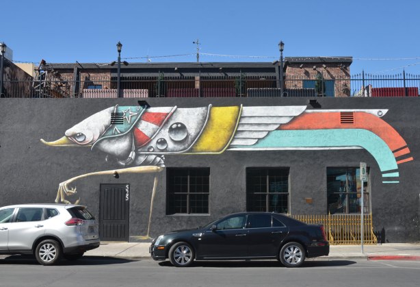 Along the front of a building there is a mural of a very big bug. It's legs are a storey high. Two cars are parked in front but the body of the bug is much higher than the cars. Bug is not realistic looking. 