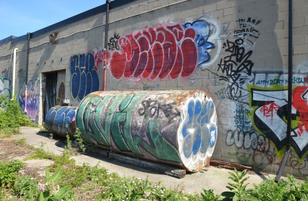 graffiti and tags on the back of a building made of concrete blocks, also on two metal storage tanks that are behind the same building. 