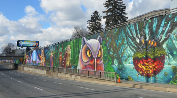 looking along a sidewalk where there is alarge mural, a very large owl is looking at you, other animals in the background.