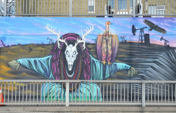 part of a larger mural on the side of an underpass, road and sidewalk in front of the photo, houses behind - a creature with a deer skull and antlers as head with a vulture sitting on its shoulder. Background scenery is dead land with an oil derrick on it.