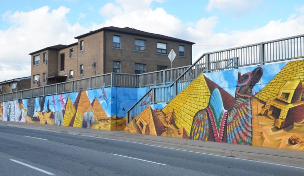 part of a larger mural on the side of an underpass, road and sidewalk in front of the photo, houses behind - looking at an angle back at the mural, Egyptian pyramids painted beside the stairs that go from the sidewalk to the houses above. Also a painted camel with a bright patterned blanket on its back.