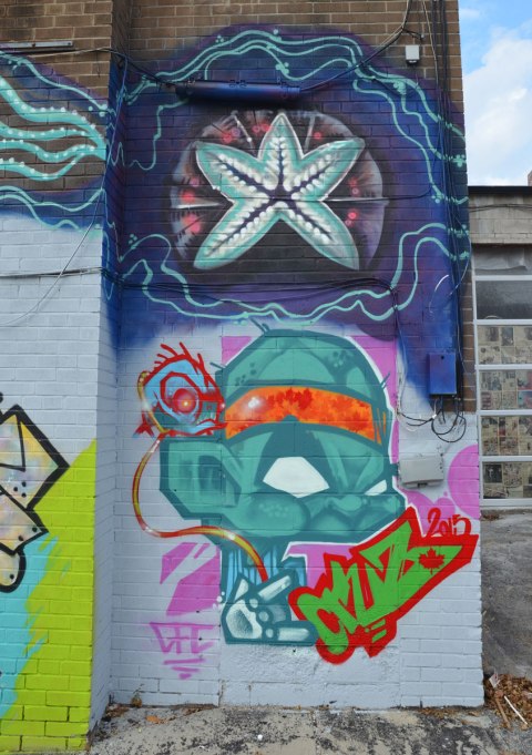 street art on the back of building. On the top is a starfish shape in blues. On the bottom is a grumpy little man with an oversized head done in teal colours. He is wearing an orange headband. GFC Cruz