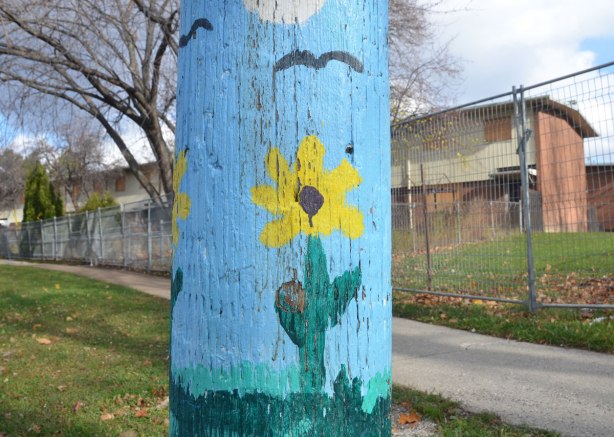 A telephone pole that has been painted on the bottom few feet. A bright blue sky with a cloud and a few birds flying, green grass and a couple of yellow flowers standing tall 