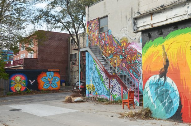 the west part of the back of a building covered with street art. The street and another lower building with a mural on it is in the background. 
