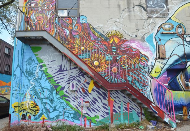 The back of a store with street art on it. A metal staircase goes diagonally up across the building. Above the stairs is a multicoloured swirly piece while under the stairs the street art in is blue and purple A campfire burns yellow in the bottom left corner.
