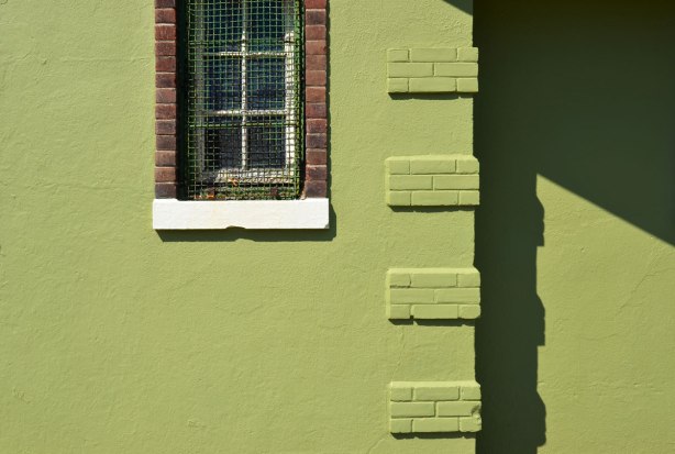 A green wall with a window taken on a sunny day when the shadows are strong. Window has brick window frame and white window sill
