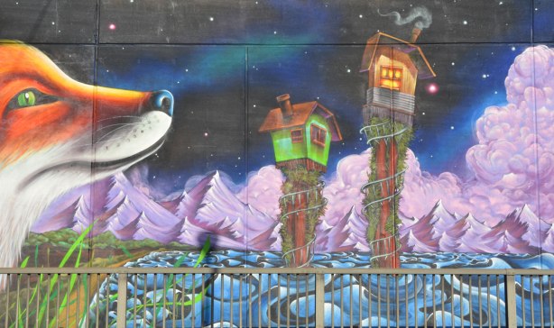 part of a mural painted on an underpass - the nose of a very large fox plus two tiny houses on stilts above turbulent water, night sky