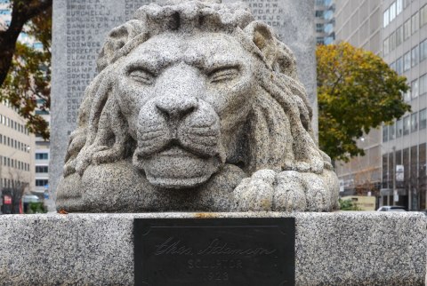 A carving of a lion in granite. It is at the base of a sculpture. A small brass plate is attached in front of the lion and it says Chas Adamson, sculptor 1923