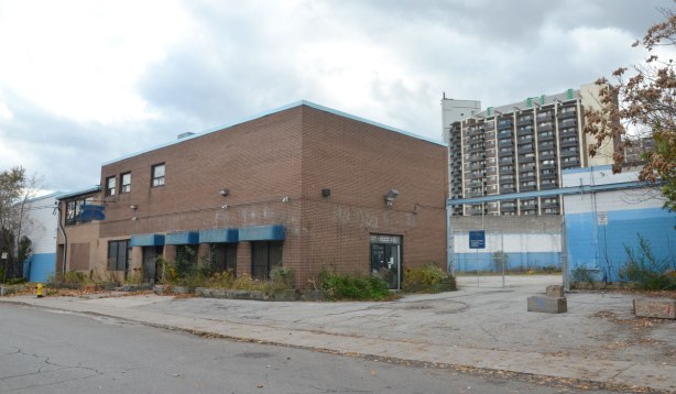 box like brick building with square awnings over the windows, large empty parking lit beside it, white and blue fence behind the parking lot, taller apartment building beyond the fence. 