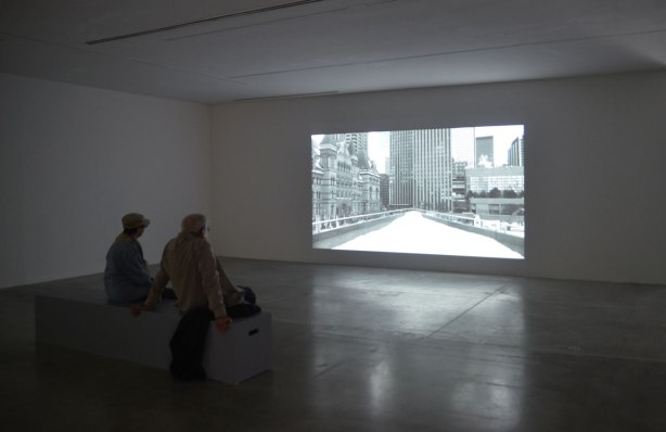 An older couple are sitting on a bench at an art gallery. They are watching a black and white film that is showing on a large screen in front of them. The image on the screen is a shot of the upper ramp at Nathan Phillips Square, looking south, in the winter with snow on the ground. There are no people in the picture on the screen. 