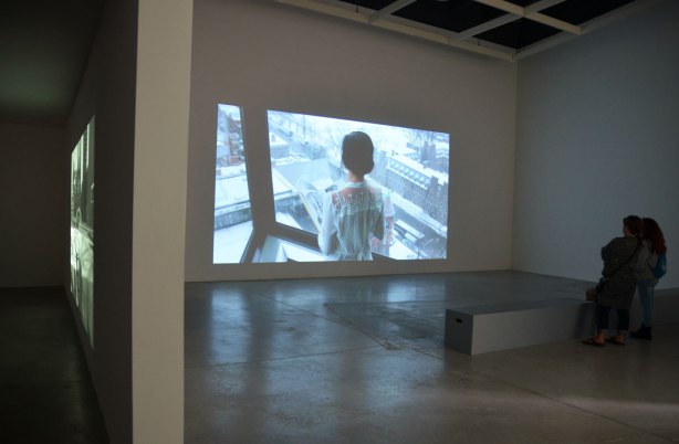 Two women are standing in the semi darkness in a room in an art gallery, watching a black and white film that is showing on a large screen in front of them. The image on the screen is the back of the upper part of a woman as she stands in front of a window in the Robarts Library in Toronto. The scene outside the window is clearly visible, winter time, University of Torotno campus. She is holding a book in her hands. 