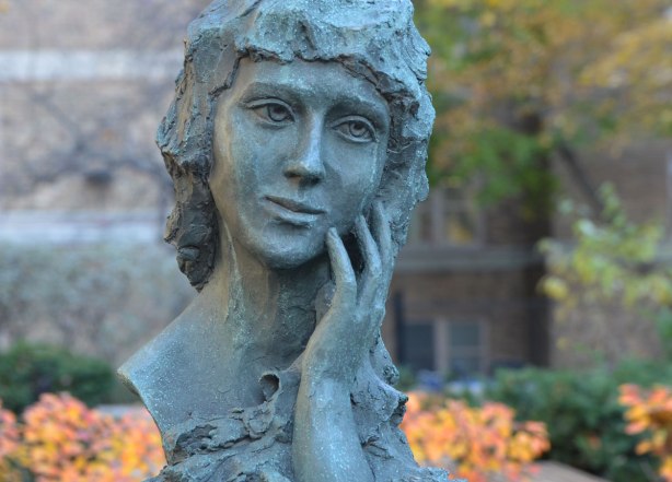 a bust of Mary Pickford, she is resting her head in one of her hands. In the background is a building along with some bushes and a tree with yellow and orange leaves.