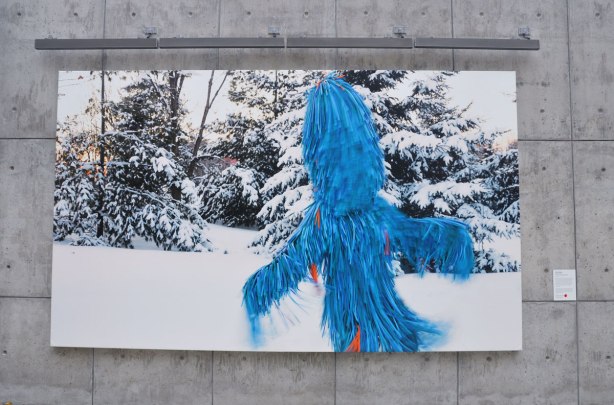 Photo of a blue furry creature taken in winter with snow covered evergreens in the background. Mounted on a concrete wall outside. 