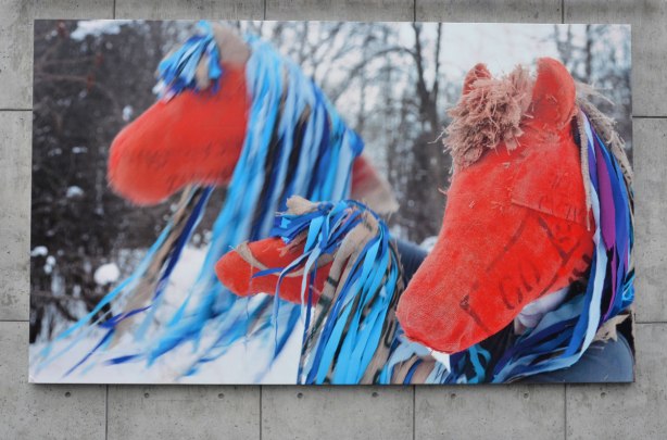 A large photograph of three red hobby horses with long blue mane, taken outside in the winter in the snow, with bare trees in the background. A mix of the real (outdoors) and the unreal (hobby horses instead of real horses). Photo is Mounted on a concrete wall outside. 
