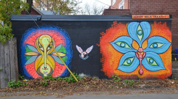 A mural by Monica on the Moon on a dark blue or black background, a bright orange and blue flower on the right and a flower/face on the left. The words Gujran Wala Tikka house are written in the top right corner which is the name of the restaurant that this building is behind. 