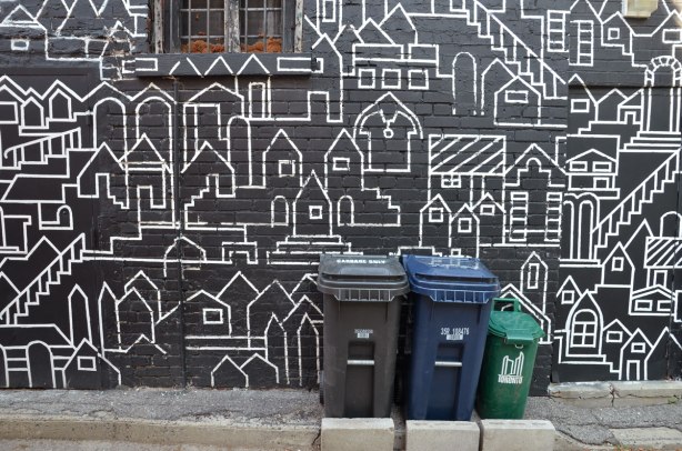 close up of part of mural, white line drawings of a city scene, lots of houses and other buildings, on a black background on the side of a house. Small window as well as three trash bins are in the picture. 
