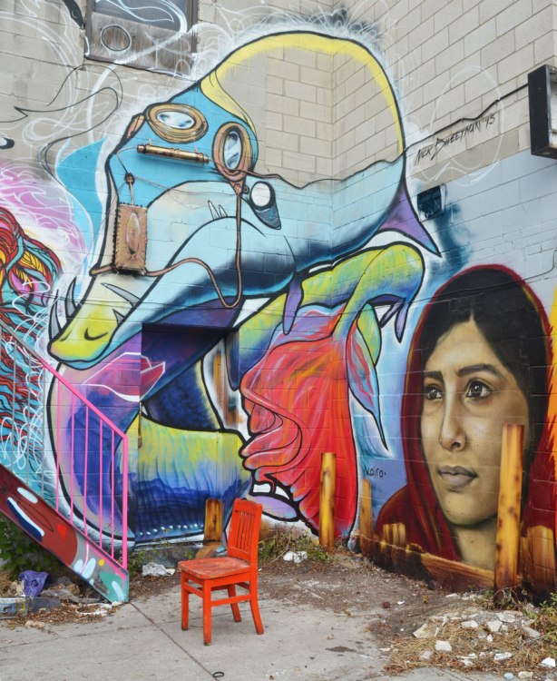 An orange chair sits in front of a corner of a building that is covered with street art. There is a realistic woman's face by kairo and a creature by Nick Sweetman