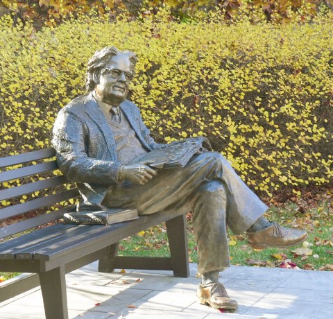 A statue of a man, Northrop Frye, sits on a bench with his legs crossed and an open book on his lap. Another book sits beside him on the bench. 