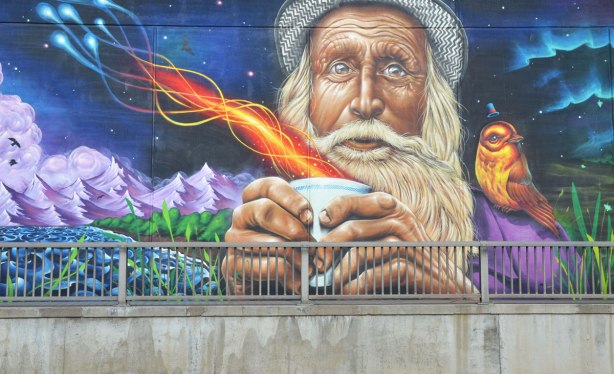 part of a large mural on an underpass in Toronto painted be Essencia Art Collective, an old man with white hair, beard and mustache and wearing a wool hat is holding a paper cup that has fire and steam coming out of it. A bird is sitting on his shoulder