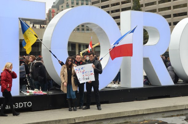 Three people standing in front of the 3D Toronto sign, holding a French flag and a large sign that says "Pas Feur, no fear"