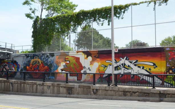 part of a large collection of street art paintings on a wall of a railway underpass on Bloor St. West as seen from across the street,
